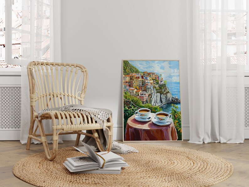 May include: A wicker chair with a throw blanket sits on a jute rug in front of a painting of a colorful Italian village on a cliff overlooking the ocean. Two cups of coffee are on a table in the painting.