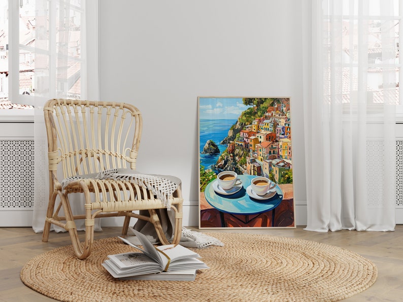 May include: A painting of a colorful Italian village on a cliff overlooking the ocean. Two white coffee cups sit on a blue table in the foreground.