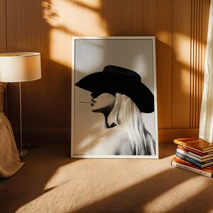 May include: A black and white framed print of a woman wearing a cowboy hat and smoking a cigarette. The artwork is leaning against a wooden wall. A stack of books sits on the floor next to the artwork. A lamp and bed are in the background.