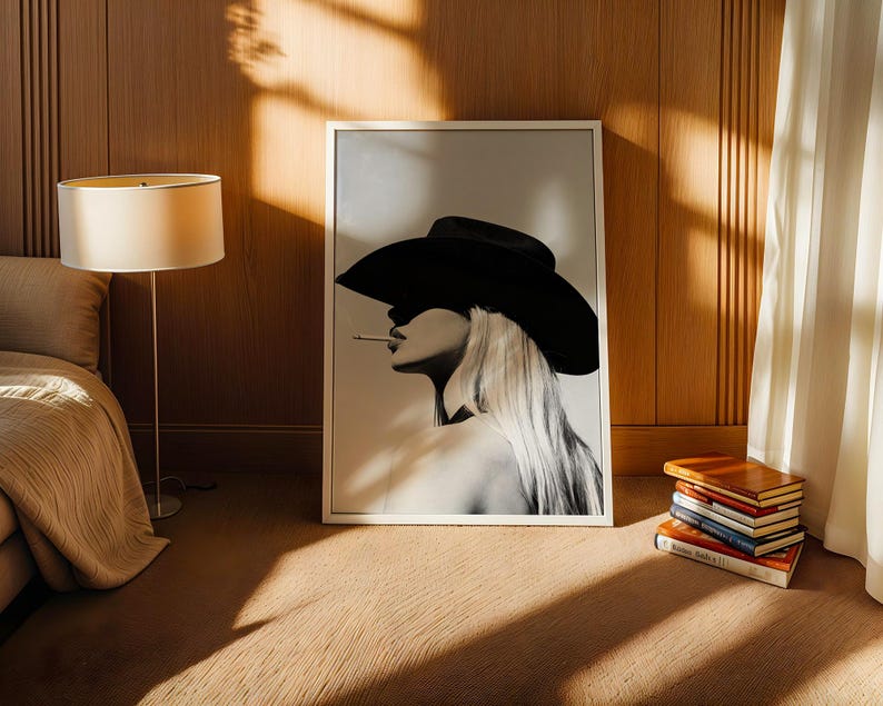 May include: A black and white framed print of a woman wearing a cowboy hat and smoking a cigarette. The artwork is leaning against a wooden wall. A stack of books sits on the floor next to the artwork. A lamp and bed are in the background.