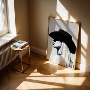 May include: A framed black and white art print of a woman wearing a large black hat and smoking a cigarette. The artwork is leaning against a wall in a room with a wooden floor. A small round table with magazines sits near a window.