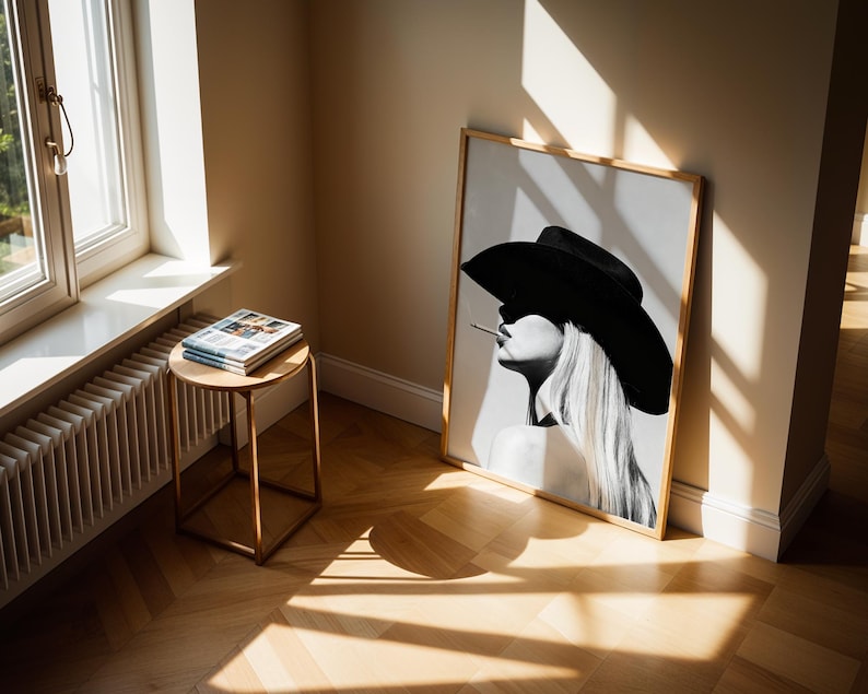 May include: A framed black and white art print of a woman wearing a large black hat and smoking a cigarette. The artwork is leaning against a wall in a room with a wooden floor. A small round table with magazines sits near a window.