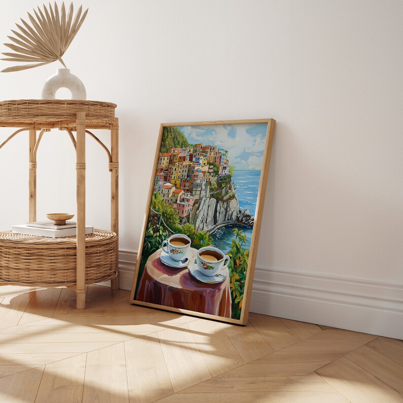 May include: A framed painting of a colorful Italian village perched on a cliff overlooking the ocean. Two cups of coffee are placed on a table in the foreground, suggesting a relaxing moment.