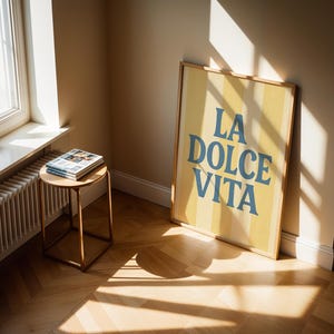 May include: A framed poster with the text "LA DOLCE VITA" in blue letters on a yellow striped background. The poster leans against a beige wall. A small round table with a stack of magazines sits near a window.
