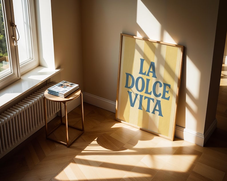 May include: A framed poster with the text "LA DOLCE VITA" in blue letters on a yellow striped background. The poster leans against a beige wall. A small round table with a stack of magazines sits near a window.