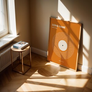May include: An orange framed print with a vinyl record design, leaning against a wall. The print features the text "SONG NAME" and "ARTIST NAME" at the top. A small, round side table with a stack of magazines sits near a window.