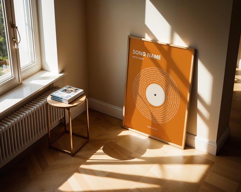May include: An orange framed print with a vinyl record design, leaning against a wall. The print features the text "SONG NAME" and "ARTIST NAME" at the top. A small, round side table with a stack of magazines sits near a window.