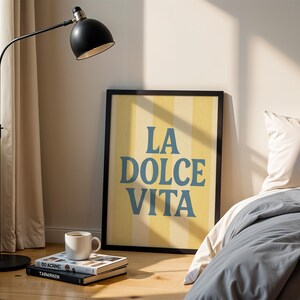 May include: A framed poster with the text "LA DOLCE VITA" in blue letters against a yellow and white striped background. A black floor lamp and a white mug on a stack of books are in the foreground.