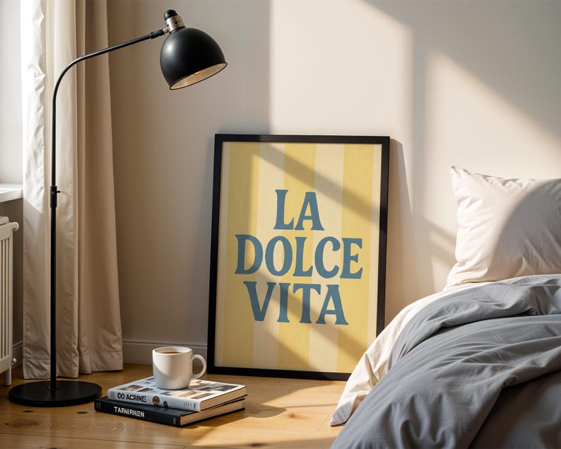 May include: A framed poster with the text "LA DOLCE VITA" in blue letters against a yellow and white striped background. A black floor lamp and a white mug on a stack of books are in the foreground.