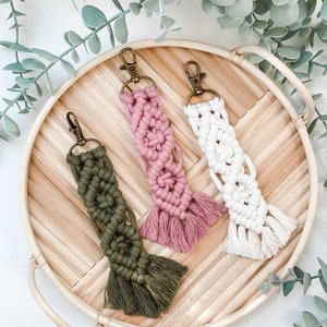 May include: Three macrame keychains with tassels in pink, white, and green hanging on a wooden tray.