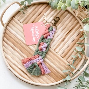 Może przedstawiać: Różowo-zielony brelok z makramą z frędzlem. Brelok ma metalowy pierścień i różową metkę z napisem "Ashley's Macrame".