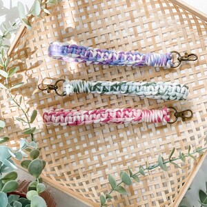 May include: Three colourful crocheted keychains with metal key rings. The keychains are in shades of purple, green, and pink. They are displayed on a woven bamboo tray.