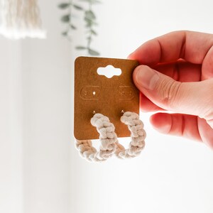 Peut inclure: Une paire de boucles d'oreilles créoles en corde blanche sur une carte marron. Les boucles d'oreilles sont faites d'une corde épaisse tressée et ont un design simple et minimaliste.