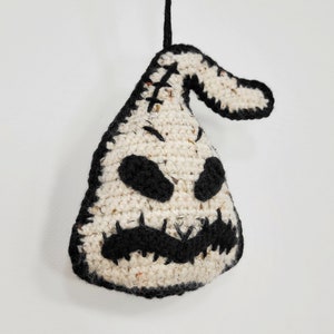 May include: A crocheted white and black ornament shaped like a ghost with a stitched-on face. The ghost has a stitched-on mouth, eyes, and a stitched-on nose. The ornament is hanging from a black string.