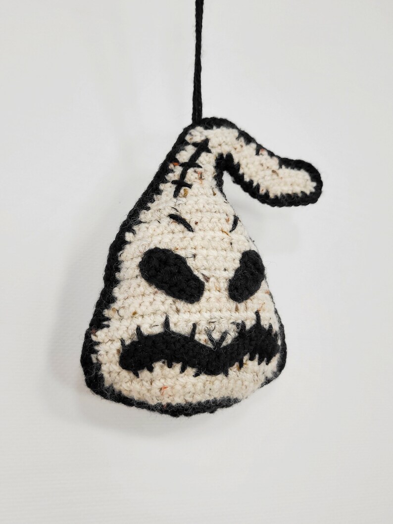 May include: A crocheted white and black ornament shaped like a ghost with a stitched-on face. The ghost has a stitched-on mouth, eyes, and a stitched-on nose. The ornament is hanging from a black string.