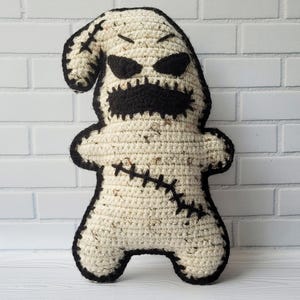 May include: A crocheted plush toy shaped like a ghost with a stitched-on black mouth, eyes, and stitches. The ghost is standing on a white wooden surface.