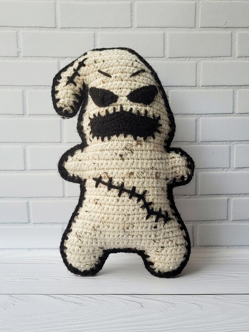 May include: A crocheted plush toy shaped like a ghost with a stitched-on black mouth, eyes, and stitches. The ghost is standing on a white wooden surface.