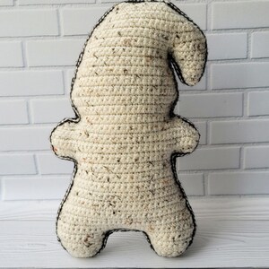 May include: A crocheted gnome doll with a cream-colored body and a black outline. The gnome is standing on a white wooden surface.
