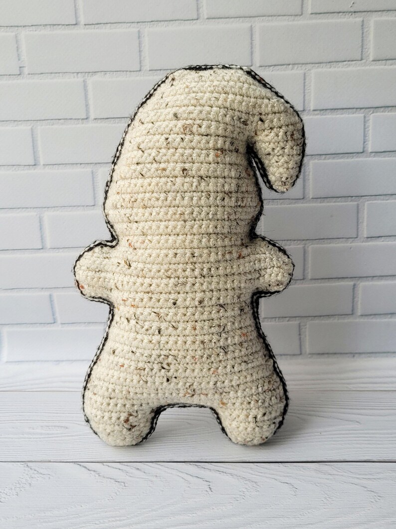 May include: A crocheted gnome doll with a cream-colored body and a black outline. The gnome is standing on a white wooden surface.