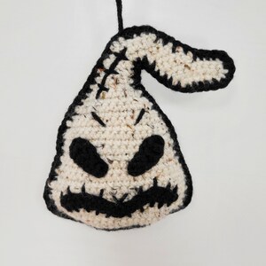 May include: A crocheted white and black ornament shaped like a ghost with a stitched-on face. The ghost has black eyes, a stitched-on mouth, and a black stitched-on nose.