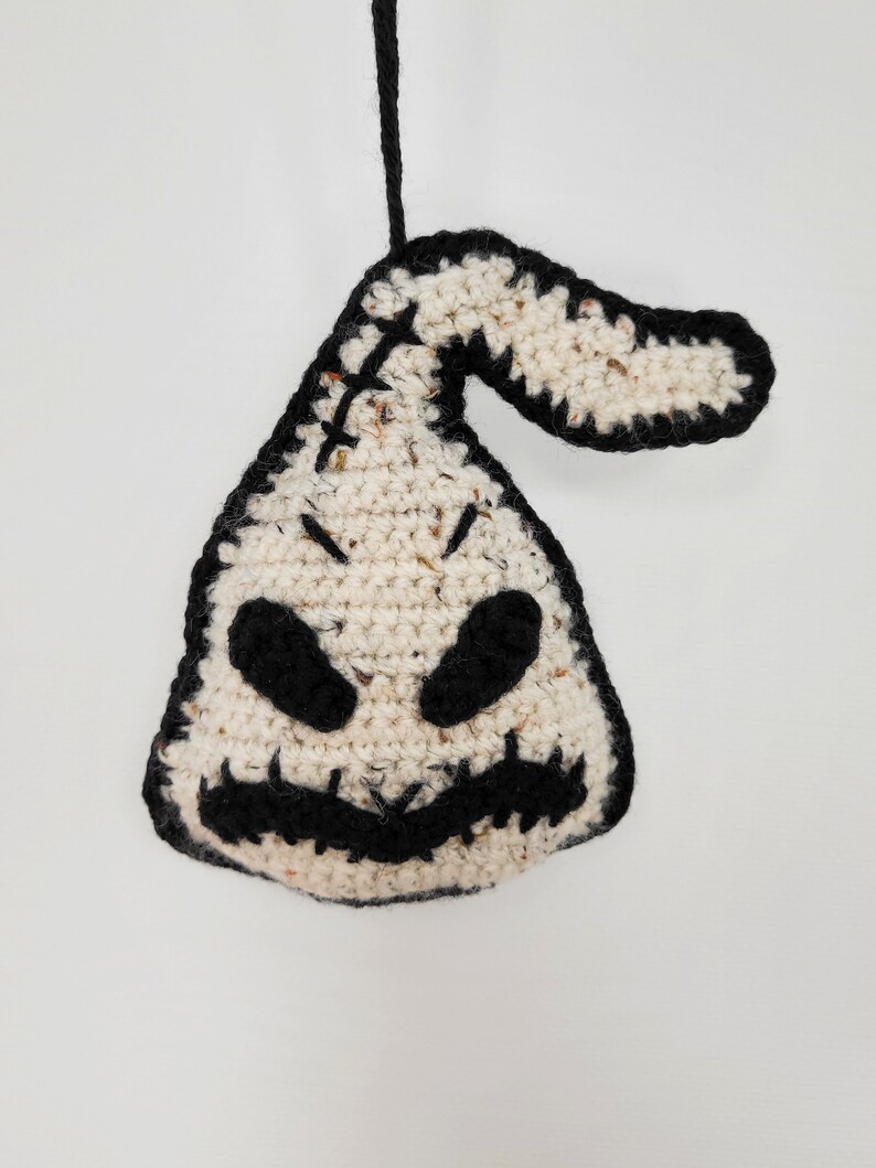 May include: A crocheted white and black ornament shaped like a ghost with a stitched-on face. The ghost has black eyes, a stitched-on mouth, and a black stitched-on nose.