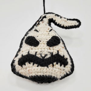 May include: A crocheted ghost ornament with a black stitched face and a black hanging loop. The ghost is white with black speckles.