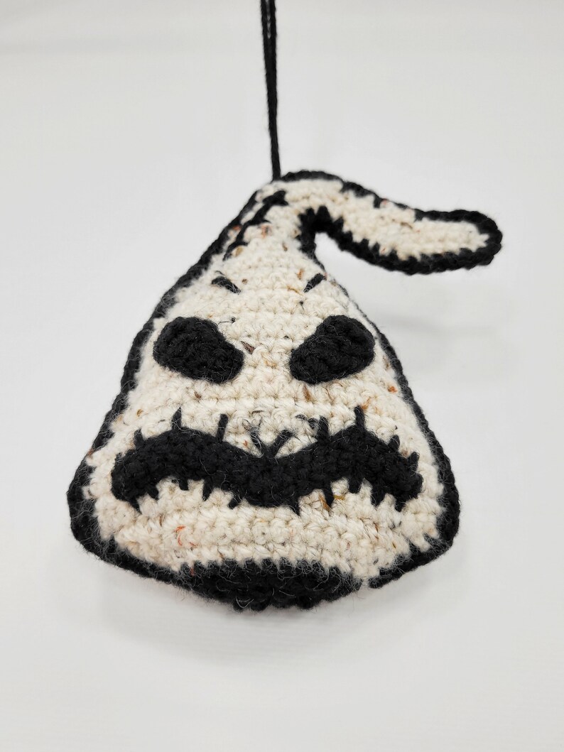 May include: A crocheted ghost ornament with a black stitched face and a black hanging loop. The ghost is white with black speckles.