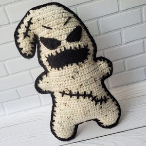 May include: A crocheted plush toy resembling the character Oogie Boogie from the movie The Nightmare Before Christmas. The toy is beige with black stitching and black facial features.