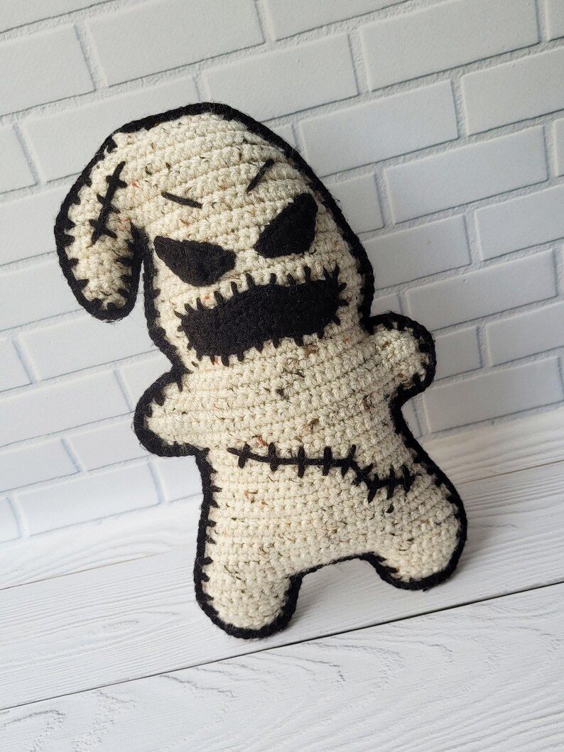 May include: A crocheted plush toy resembling the character Oogie Boogie from the movie The Nightmare Before Christmas. The toy is beige with black stitching and black facial features.