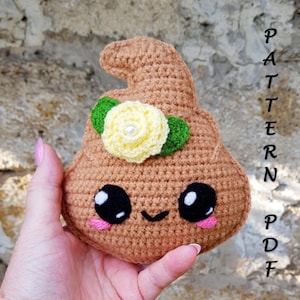May include: A crocheted brown plushie shaped like a poop emoji, featuring a smiling face with black eyes and pink cheeks. It is adorned with a yellow flower and green leaves. The text "PATTERN PDF" is written vertically on the right side.