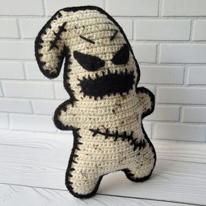 May include: A crocheted plush toy resembling a ghost with black stitched details, including eyes, a mouth, and stitches on the body. The toy is standing on a white wooden surface.