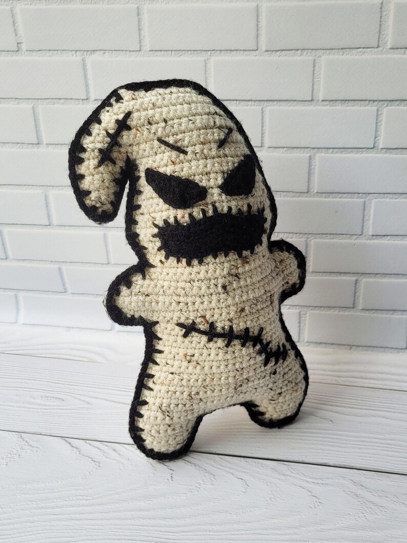 May include: A crocheted plush toy resembling a ghost with black stitched details, including eyes, a mouth, and stitches on the body. The toy is standing on a white wooden surface.