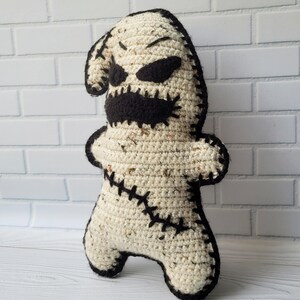May include: A crocheted plush toy resembling a stitched-up ghost with a black stitched-up mouth and eyes. The toy is standing on a white wooden surface.