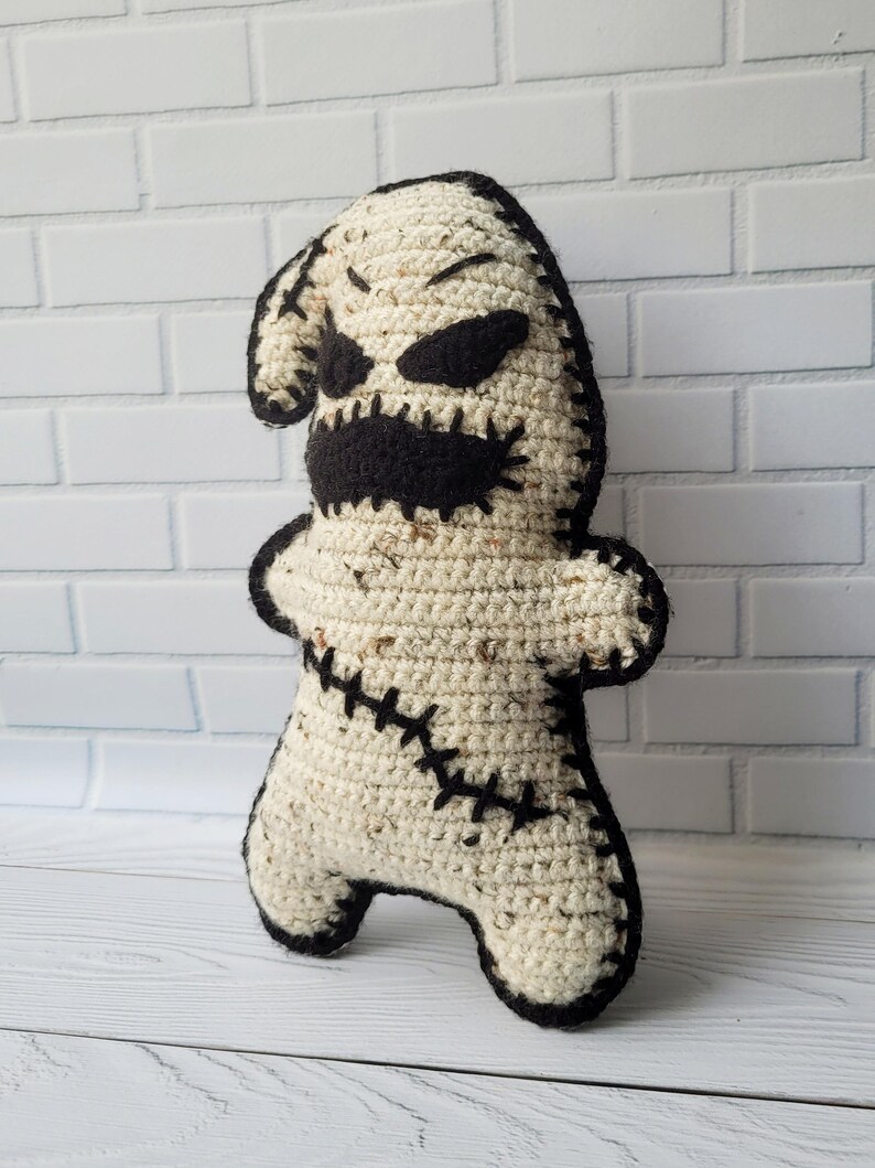 May include: A crocheted plush toy resembling a stitched-up ghost with a black stitched-up mouth and eyes. The toy is standing on a white wooden surface.