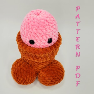 Puede incluir: Una escultura de ganchillo de felpa hecha a mano con forma de pene. La cabeza es rosa con ojos de botón negros, y el tronco y los testículos son de color naranja óxido. El texto "PATTERN PDF" está escrito verticalmente en rosa.