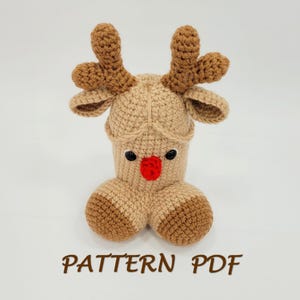 May include: A crocheted reindeer figure in tan and brown, with a red nose and black eyes. The reindeer has large antlers and a rounded body. The text "PATTERN PDF" is displayed in brown at the bottom of the image.