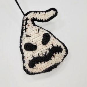 May include: A crocheted white ghost ornament with black details, including eyes, a mouth, and a stitched-on loop for hanging.