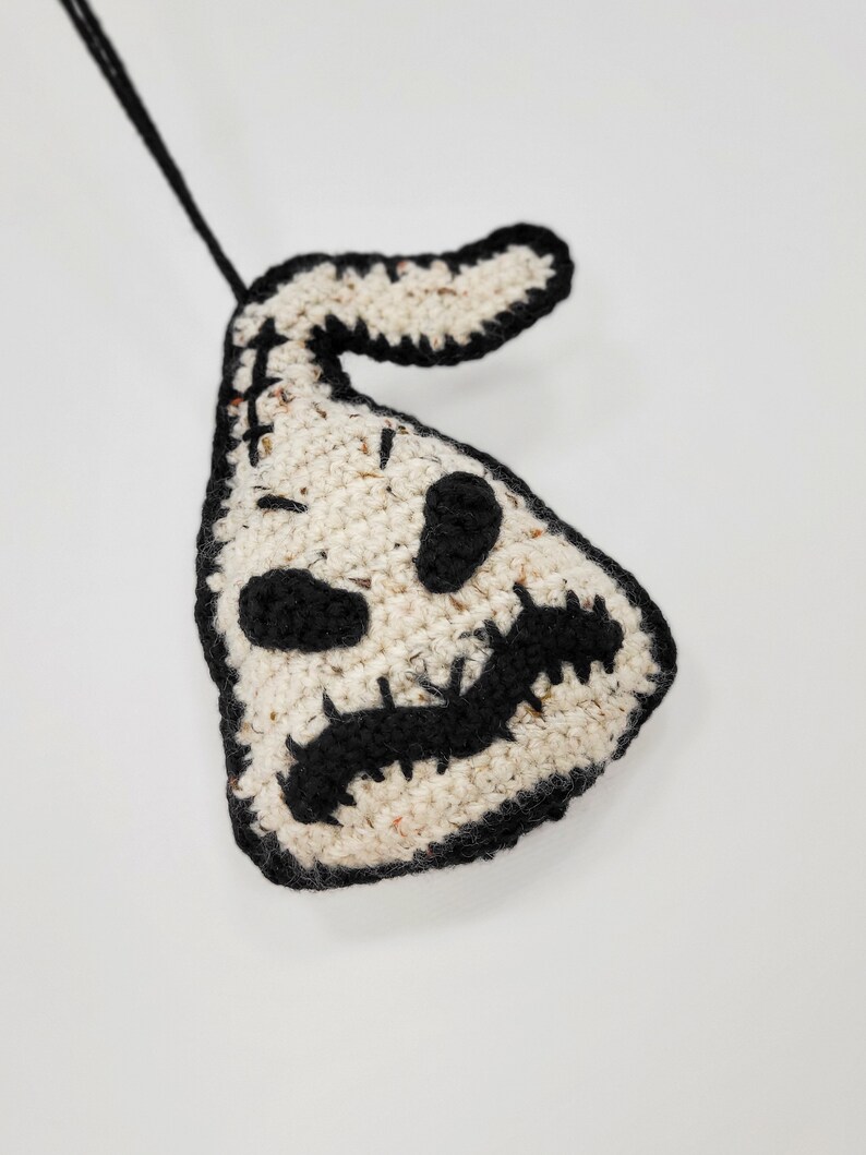 May include: A crocheted white ghost ornament with black details, including eyes, a mouth, and a stitched-on loop for hanging.