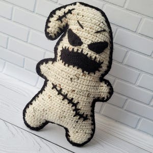 May include: A crocheted ghost plush toy with black eyes, a stitched mouth, and black stitching around the edges. The toy is standing on a white wooden surface.