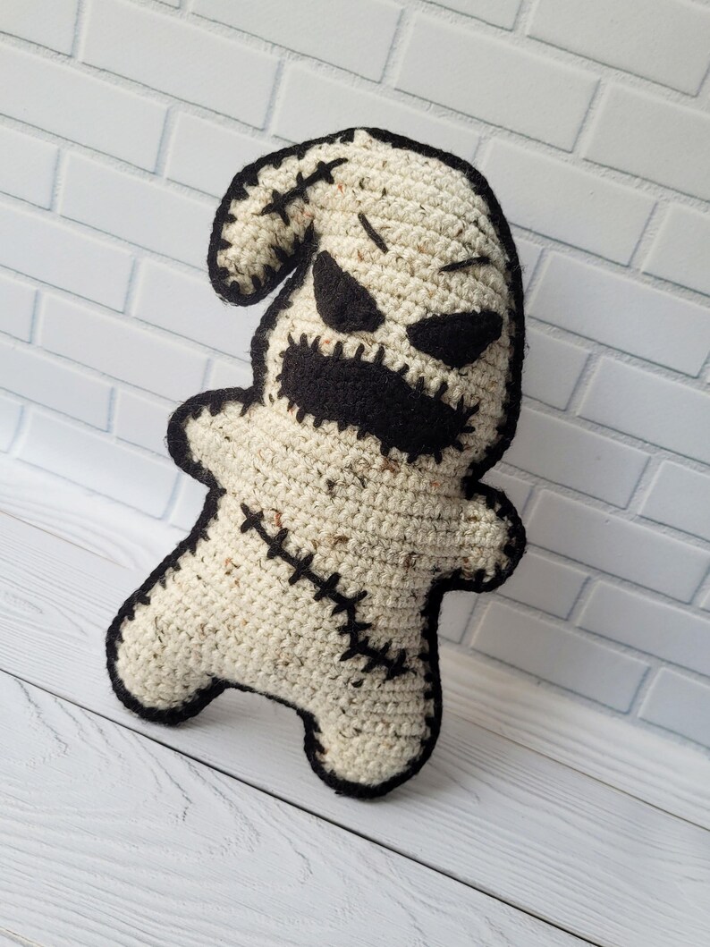 May include: A crocheted ghost plush toy with black eyes, a stitched mouth, and black stitching around the edges. The toy is standing on a white wooden surface.