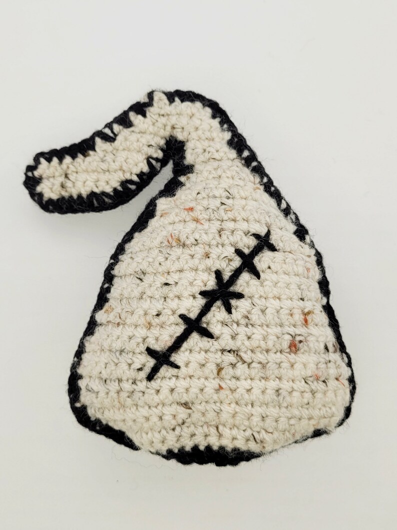 May include: A white crocheted stuffed toy shaped like a sack with a black crocheted border and a black stitched X across the front.