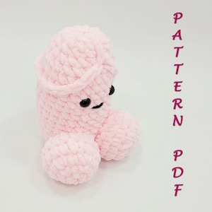 May include: A light pink, plush, crocheted phallus-shaped toy with black button eyes and a smiling mouth. The toy has two round testicles and a curved head. The word "PATTERN PDF" is written vertically in pink.