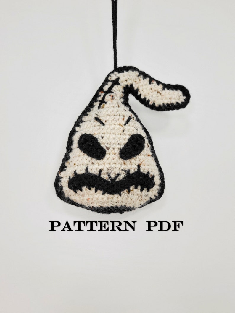 May include: Crochet pattern for a white and black ghost ornament with a stitched-on mouth and eyes. The pattern is available as a PDF download. The text "PATTERN PDF" is printed below the ornament.