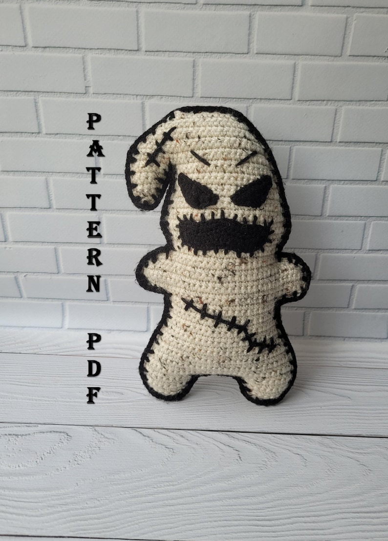 May include: A crocheted ghost amigurumi toy with black eyes, a stitched mouth, and black stitching details. The toy is standing on a white wooden surface.