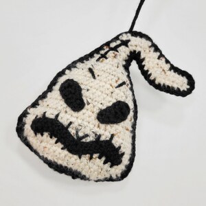 May include: A crocheted ornament shaped like a ghost with a black stitched face and a black stitched smile. The ornament is white with black accents and has a loop for hanging.