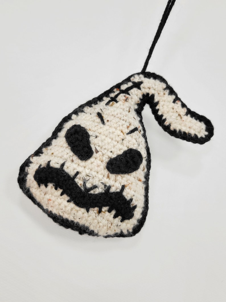 May include: A crocheted ornament shaped like a ghost with a black stitched face and a black stitched smile. The ornament is white with black accents and has a loop for hanging.