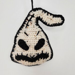 May include: A crocheted white and black ornament shaped like a ghost with a stitched-on mouth and eyes. The ornament is hanging from a black string.