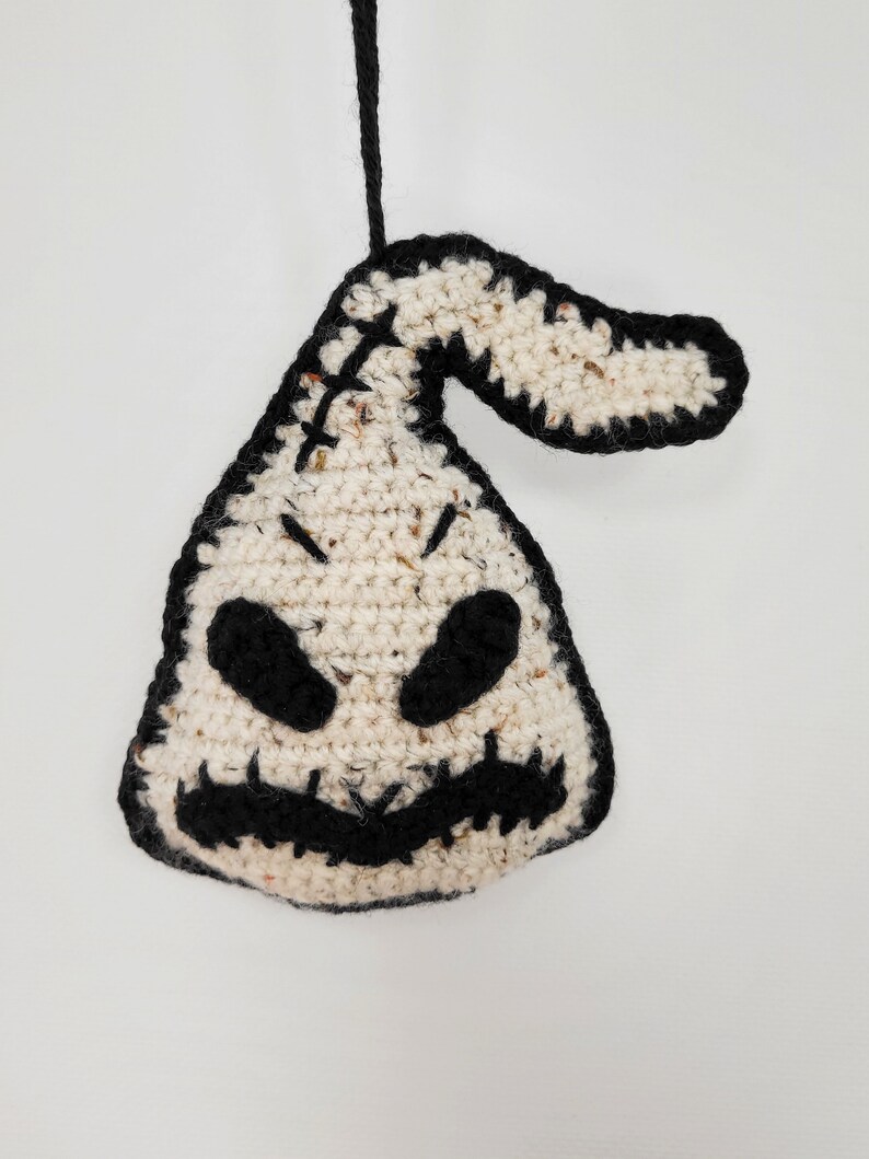 May include: A crocheted white and black ornament shaped like a ghost with a stitched-on mouth and eyes. The ornament is hanging from a black string.