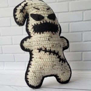May include: A crocheted plush toy in the shape of a ghost with a stitched-on face. The ghost is white with black eyes, a mouth, and stitching. The toy is standing on a white wooden surface.