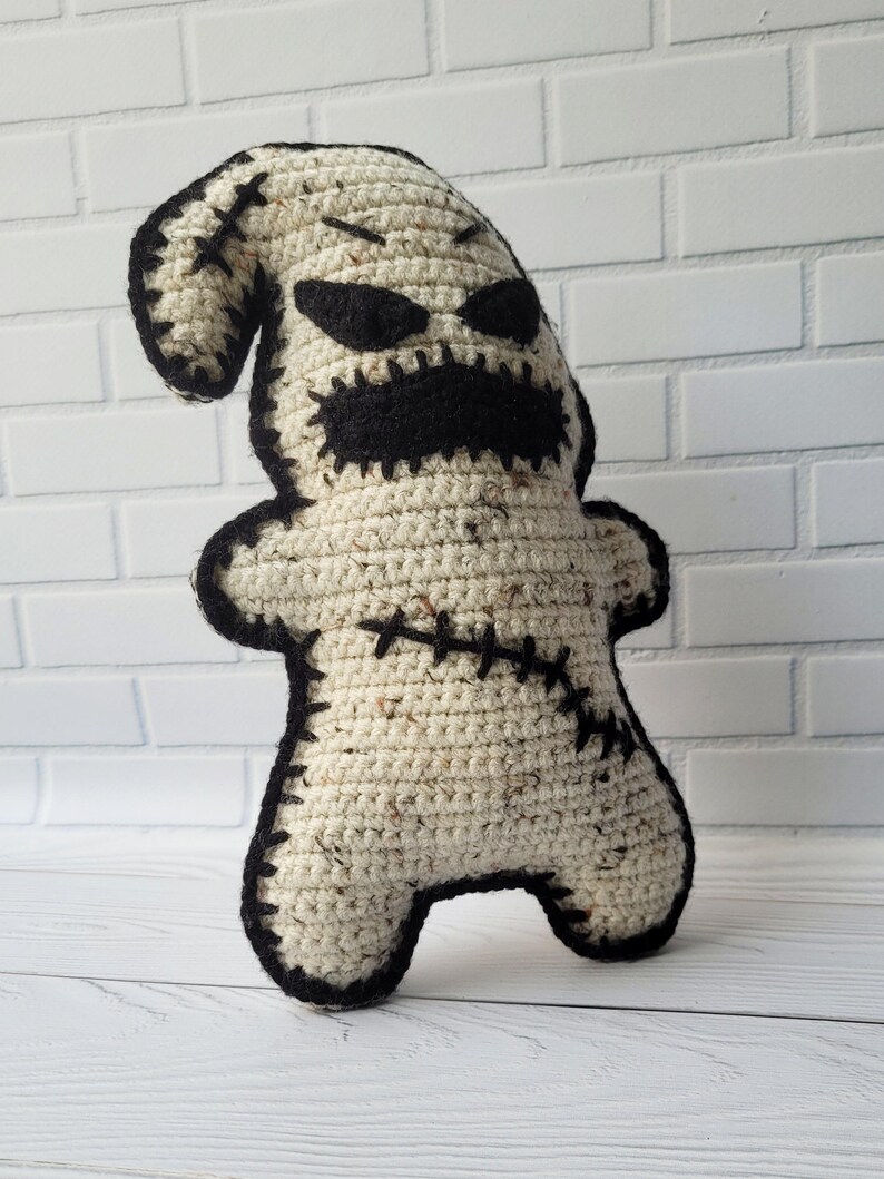 May include: A crocheted plush toy in the shape of a ghost with a stitched-on face. The ghost is white with black eyes, a mouth, and stitching. The toy is standing on a white wooden surface.